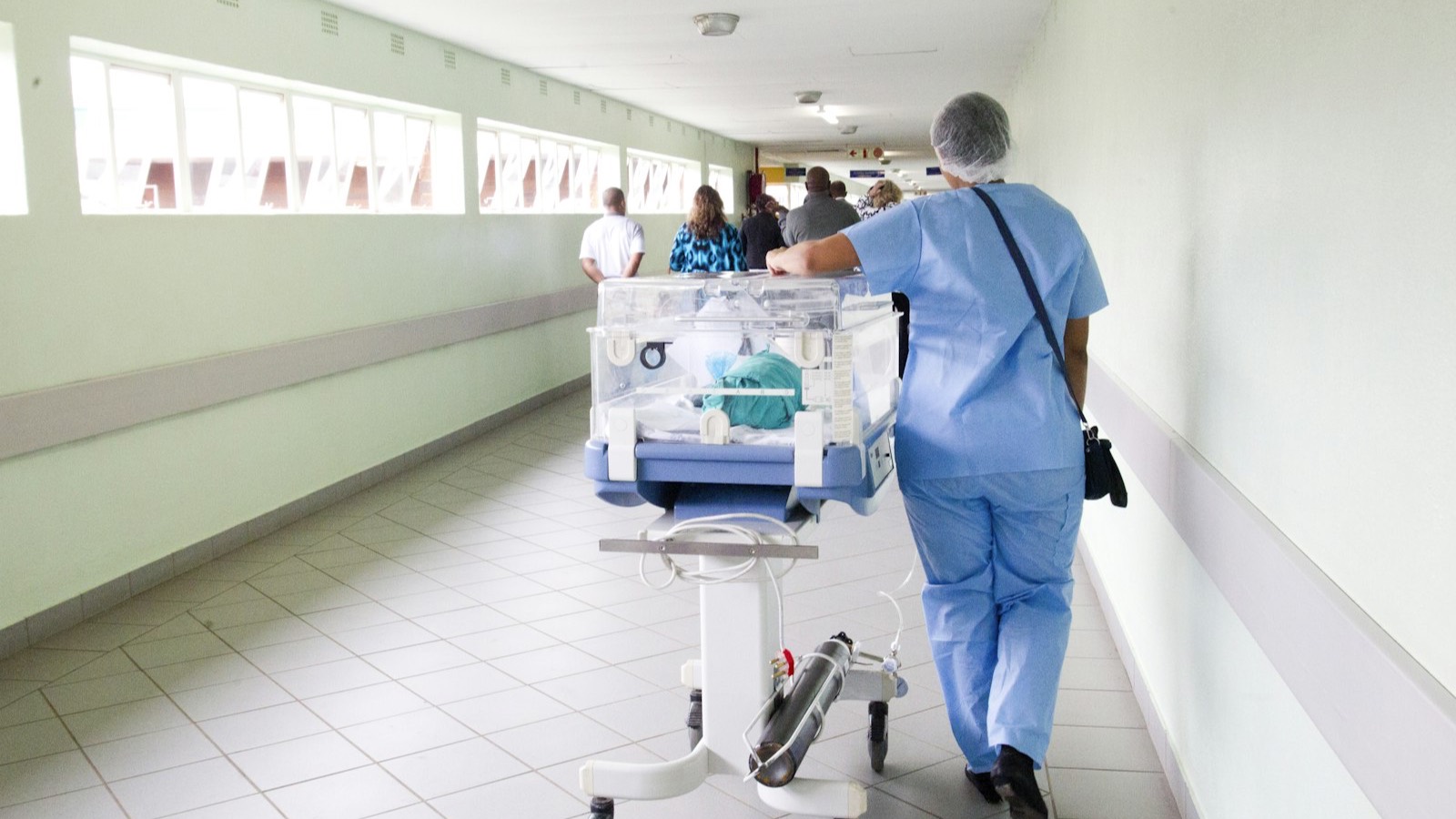A clinician in scrubs walks a hospital corridor past an incubator.